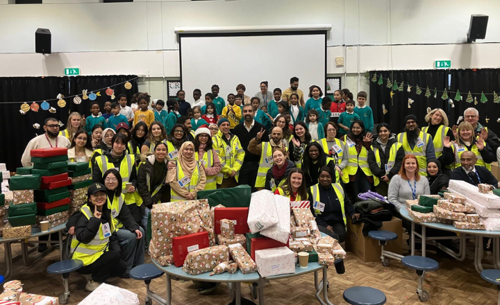 A group of people in yellow safety vests gathers around a table filled with colorful presents, smiling and chatting.