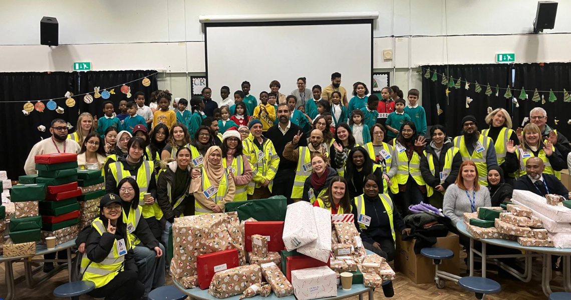 A group of people in yellow safety vests gathers around a table filled with colorful presents, smiling and chatting.