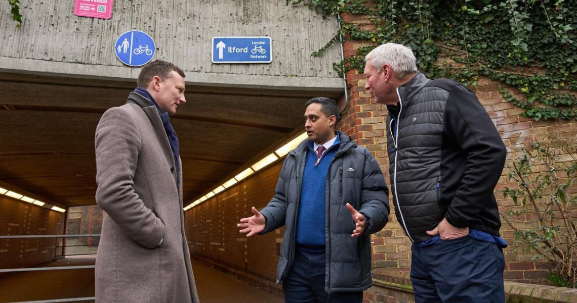  Three men engaged in conversation inside a tunnel located beneath a bridge, surrounded by concrete walls and dim lighting.