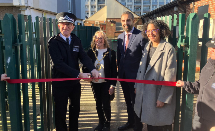 Five people at a ribbon-cutting ceremony outdoors, with a police officer holding large scissors. They are standing in front of green metal fences.