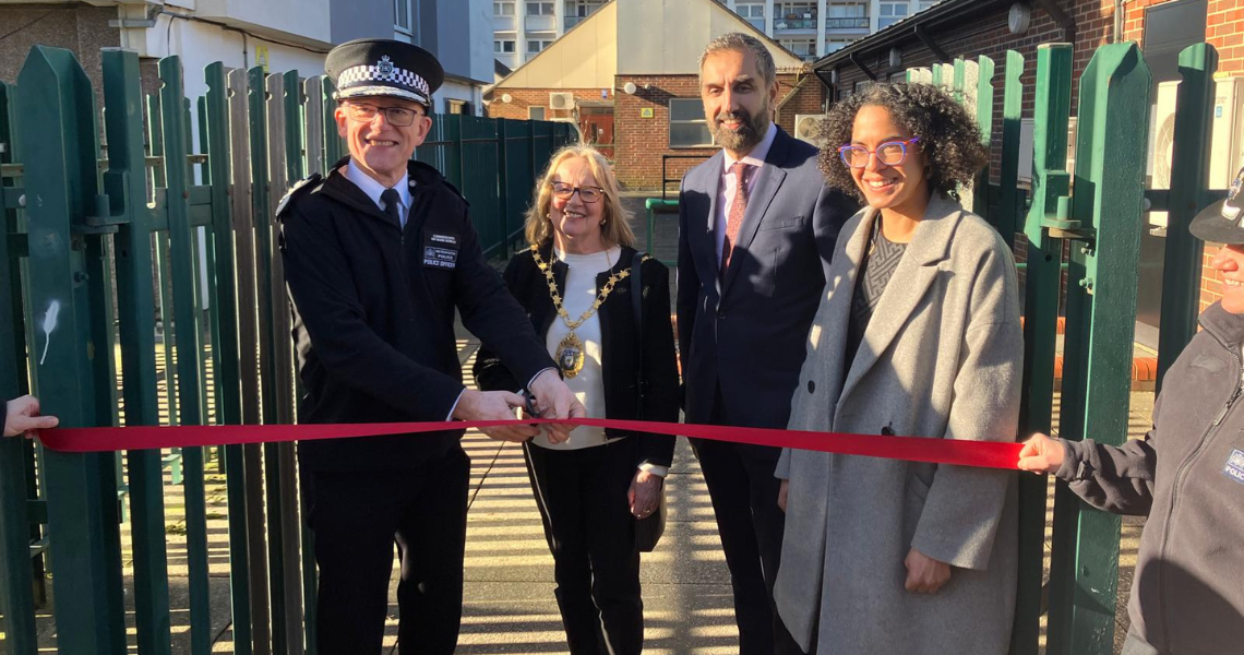 Five people at a ribbon-cutting ceremony outdoors, with a police officer holding large scissors. They are standing in front of green metal fences.