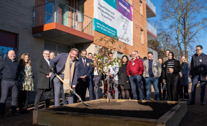 A gathering of people poses in front of a building, with a tree visible in the foreground of the scene.