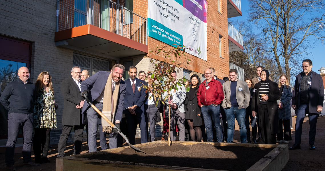 A gathering of people poses in front of a building, with a tree visible in the foreground of the scene.