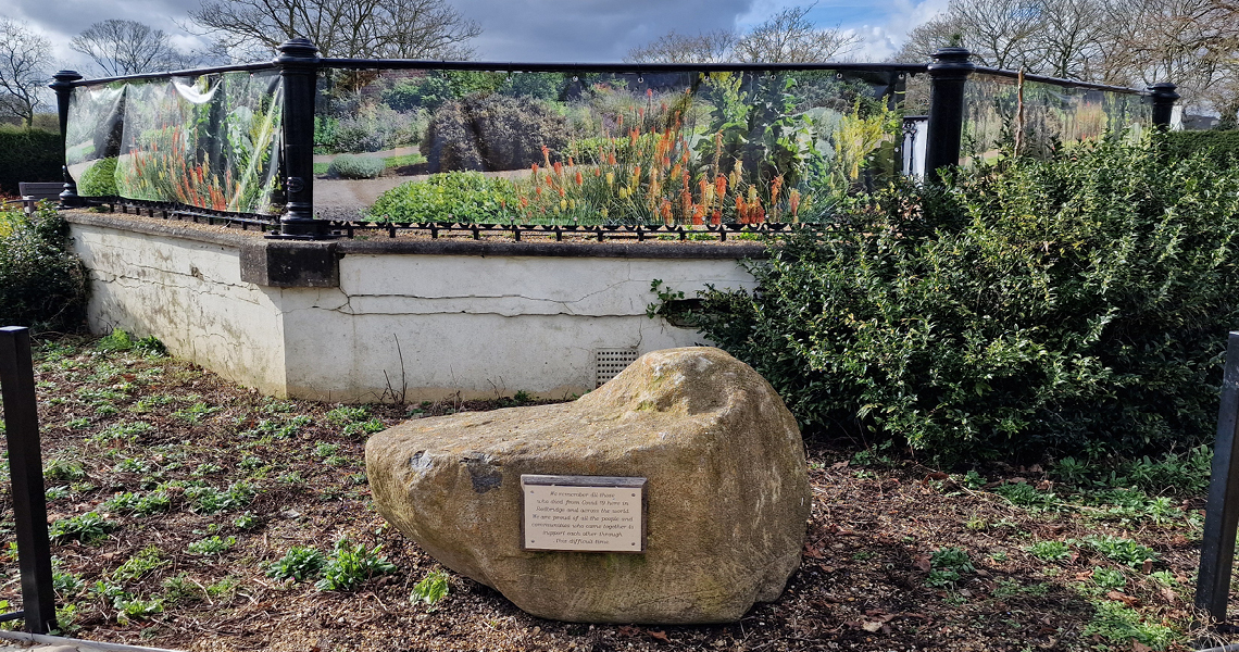 A large stone with an attached plaque is situated in front of a raised flowerbed surrounded by a printed garden image on a fence. Trees and a cloudy sky are visible in the background.